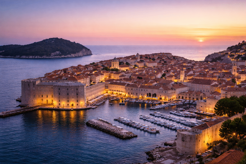 Croatia by Yacht: Aerial sunset view of Dubrovnik’s Old Town enclosed by stone walls, with boats docked in the glowing harbor along the Adriatic Sea.