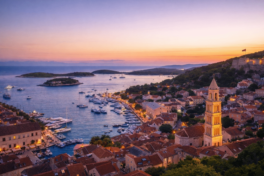 Croatia by Yacht: Sunset panorama of Hvar Town marina with sailboats and yachts anchored beside red-roofed buildings and nearby islands.
