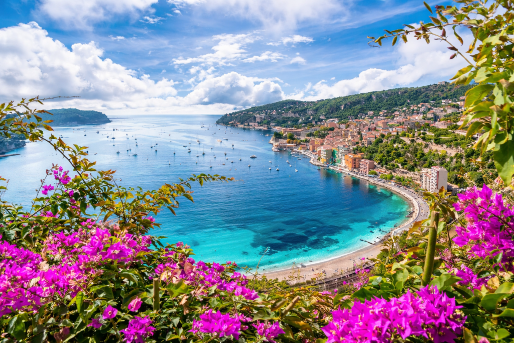 Panoramic coastal view of a turquoise bay framed by flowers in the South of France with boats scattered across calm water