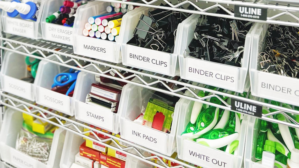 Organizing office supplies in labeled bins, including binder clips, markers, scissors, and ink pads on metal shelving.”