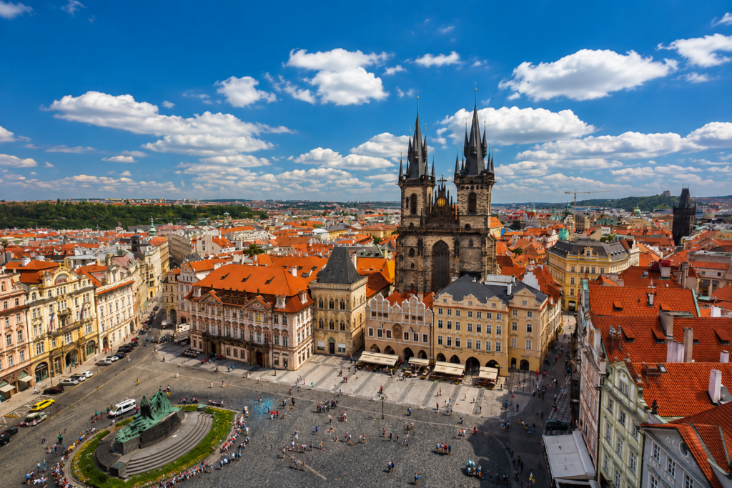 Panoramic view of Prague's Old Town Square with the Church of Our Lady before Týn, historic buildings, and red rooftops under a bright blue sky.