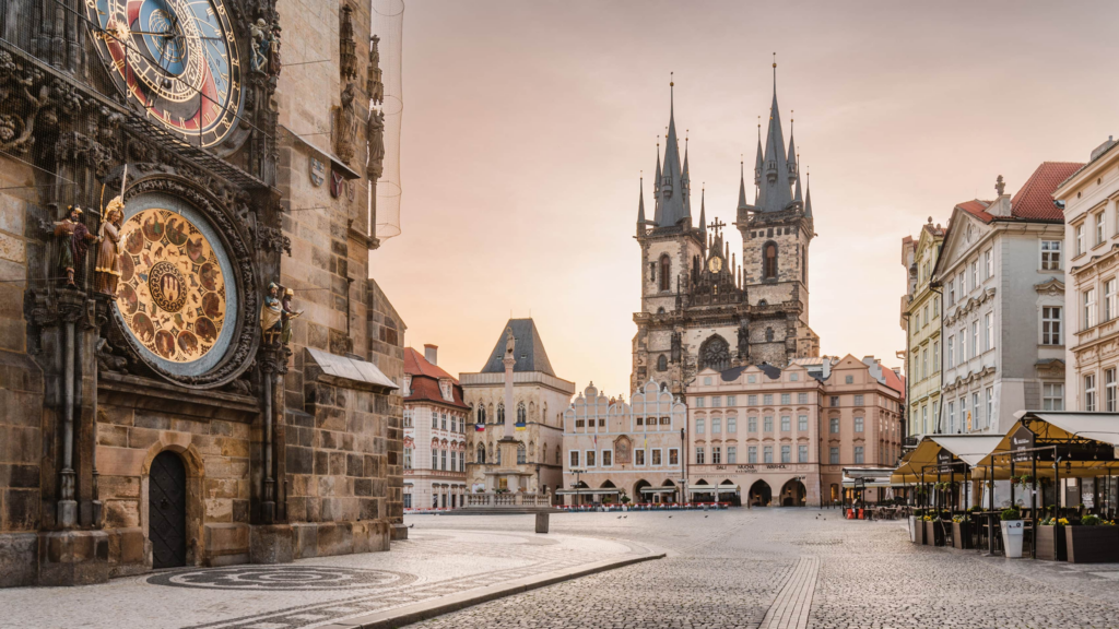 Prague's Old Town Square at sunrise, featuring the Astronomical Clock on the Old Town Hall and the Gothic towers of the Church of Our Lady before Týn, with cobblestone streets and historic buildings bathed in soft morning light.
